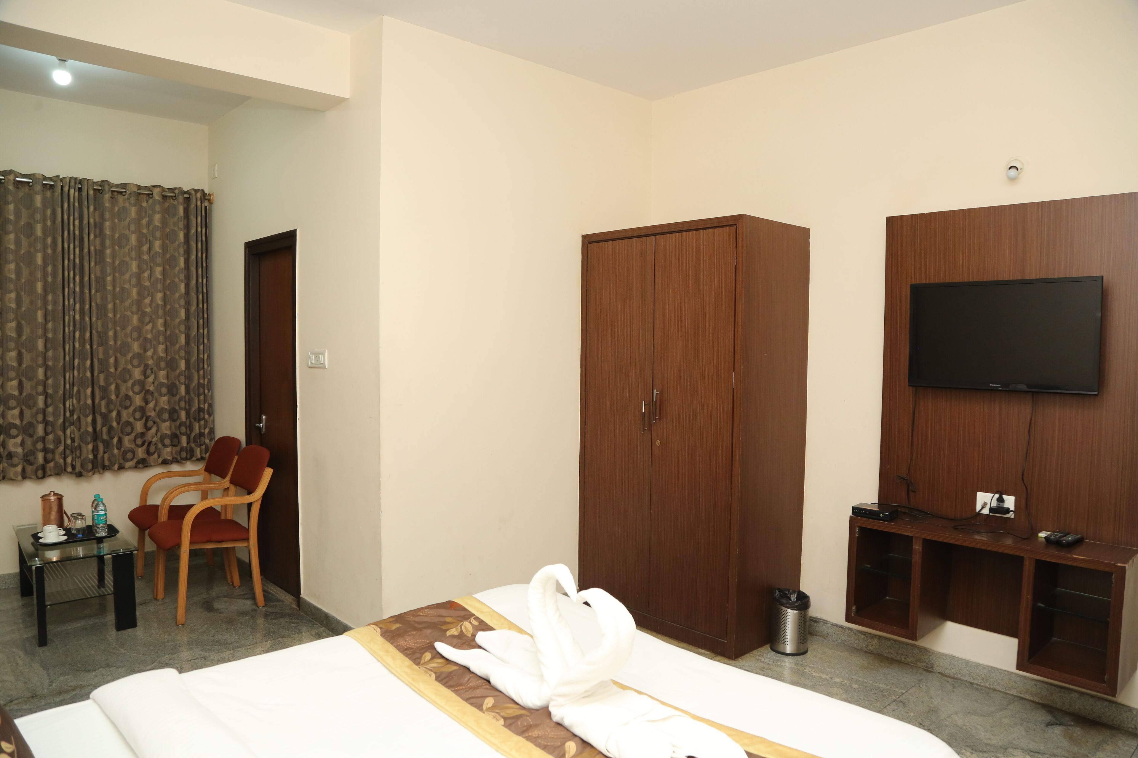 A hotel room with a bed, a TV mounted on the wall, a wardrobe, a desk with a chair, and a curtain.