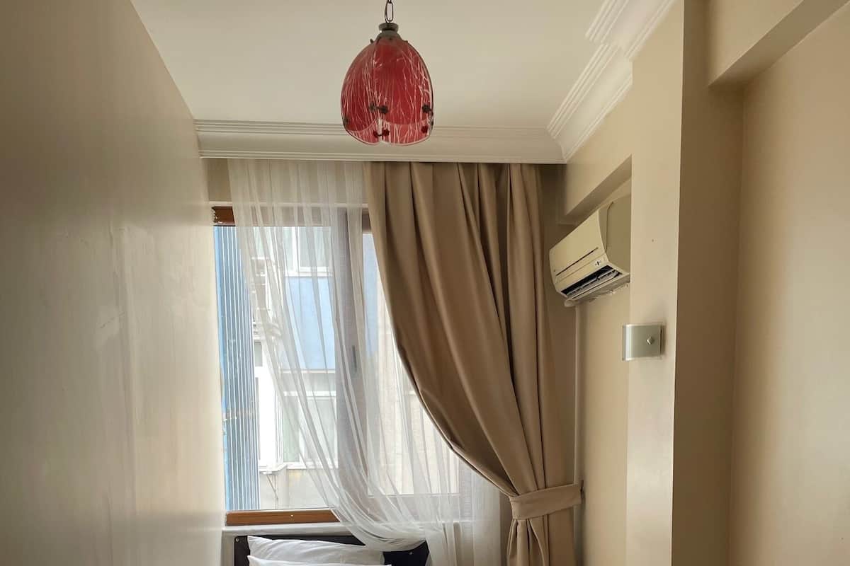 A neatly made bed with white linens, a red pendant light, and a window with sheer curtains.