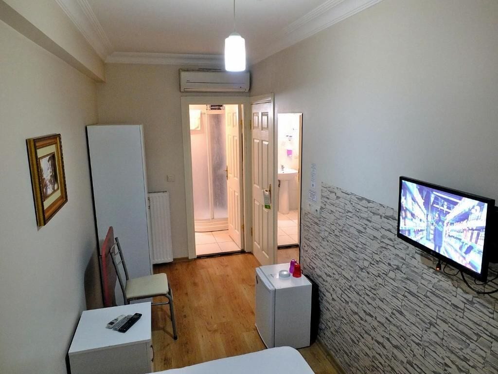 A small room with a bed, a desk, a chair, a television, and a refrigerator. There is a door leading to a bathroom.