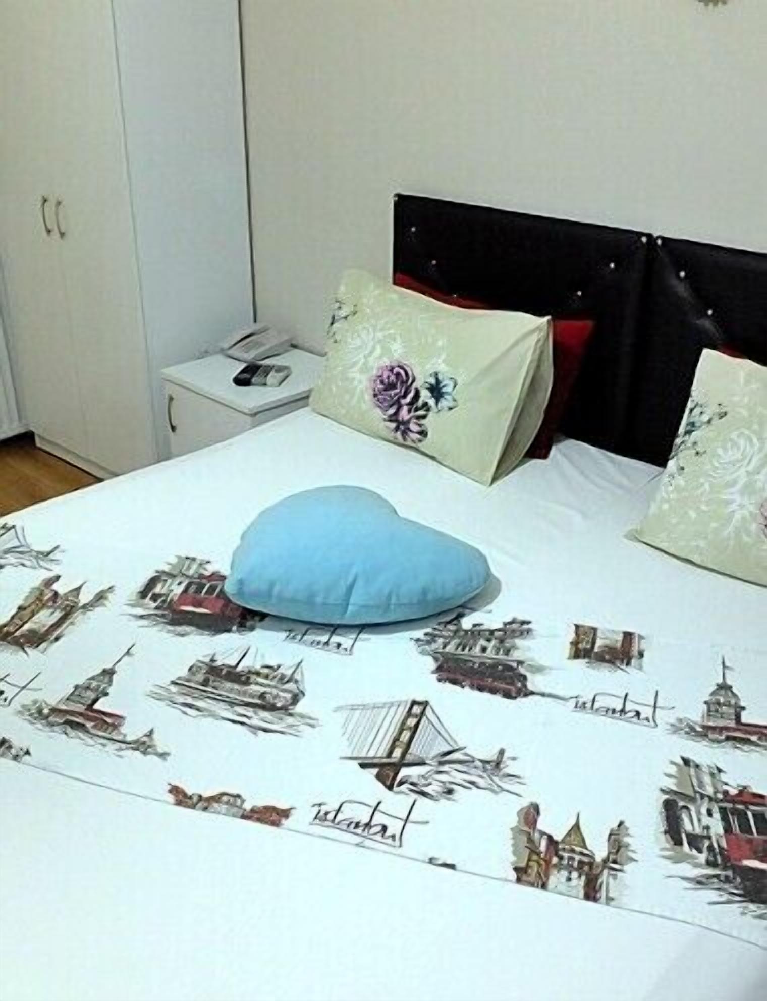 A bed with a white sheet featuring cityscape prints, a blue heart-shaped pillow, and two floral pillows.