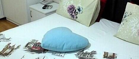 A bed with a white sheet featuring cityscape prints, a blue heart-shaped pillow, and two floral pillows.