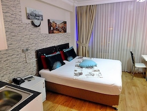 A bedroom with a bed, a small desk, a chair, a sink, and a window with curtains.