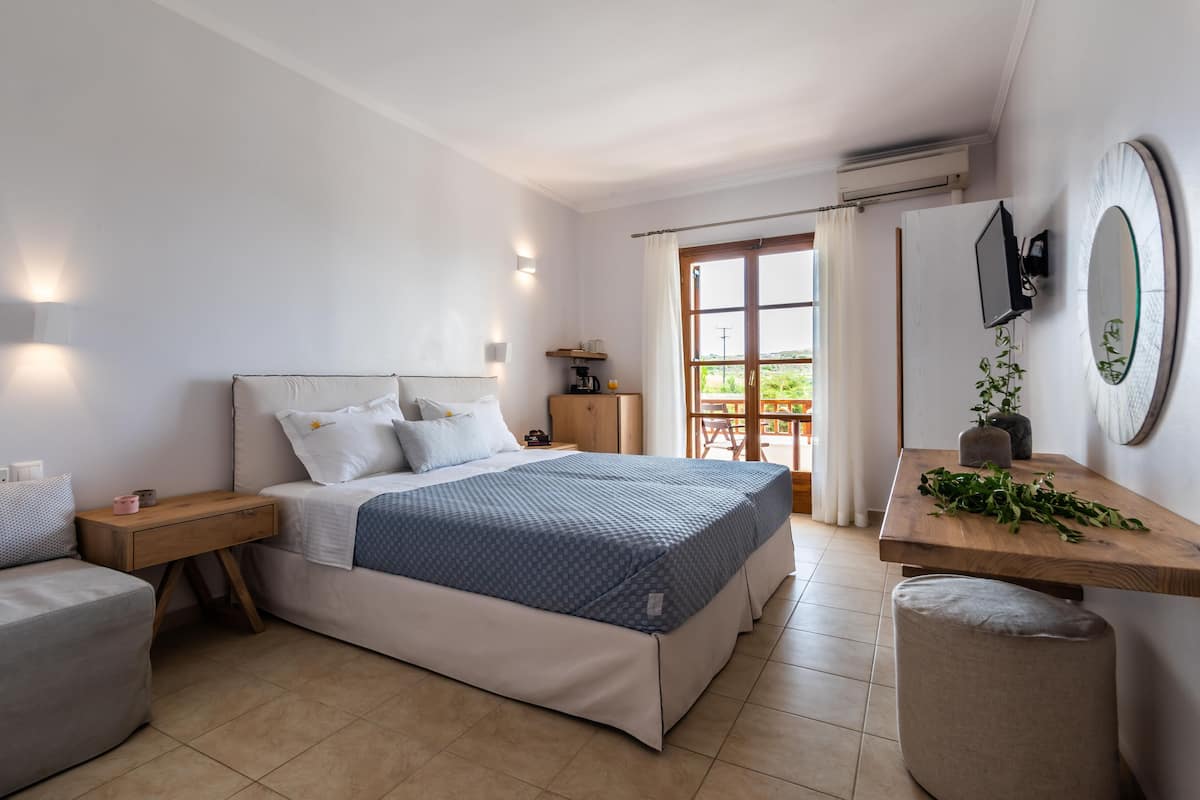 A modern bedroom with a large bed, bedside tables, a small seating area, a wooden side table, and a view of the outdoors.