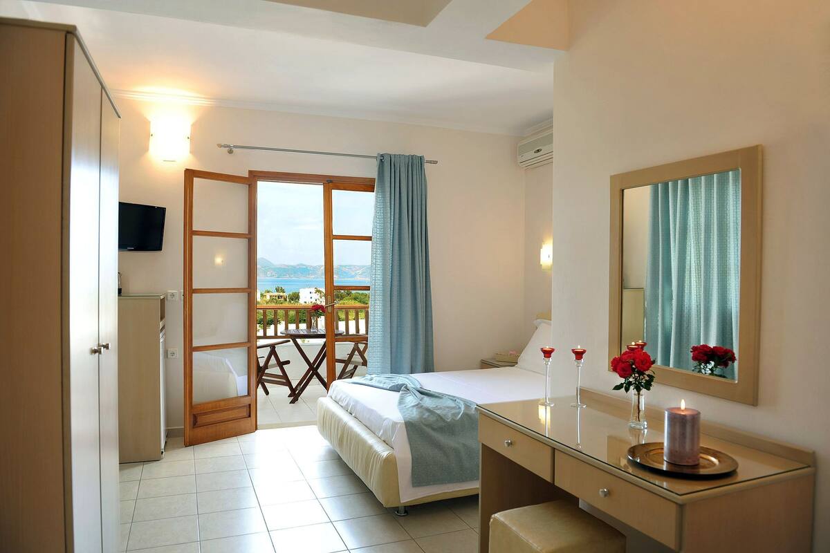 A bedroom with a bed, a bedside table with a mirror, a dresser, and a balcony with a view.
