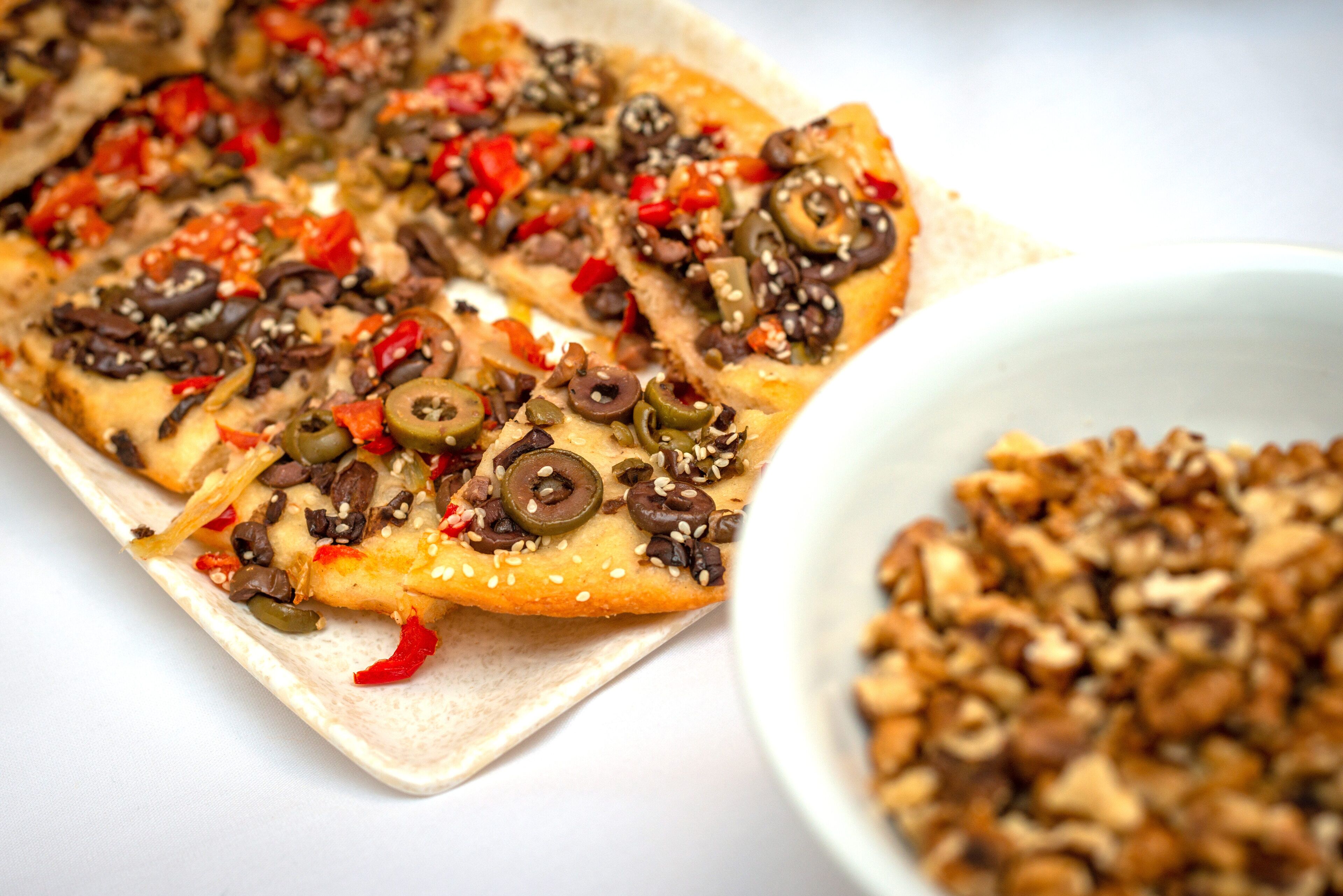 A pizza with various toppings and a bowl of nuts on a white surface.