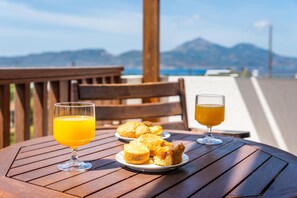 A wooden table with a glass of orange juice and a glass of beer, and two plates of biscuits on a terrace with a view of the sea and mountains. - Konstantinos (Milos)