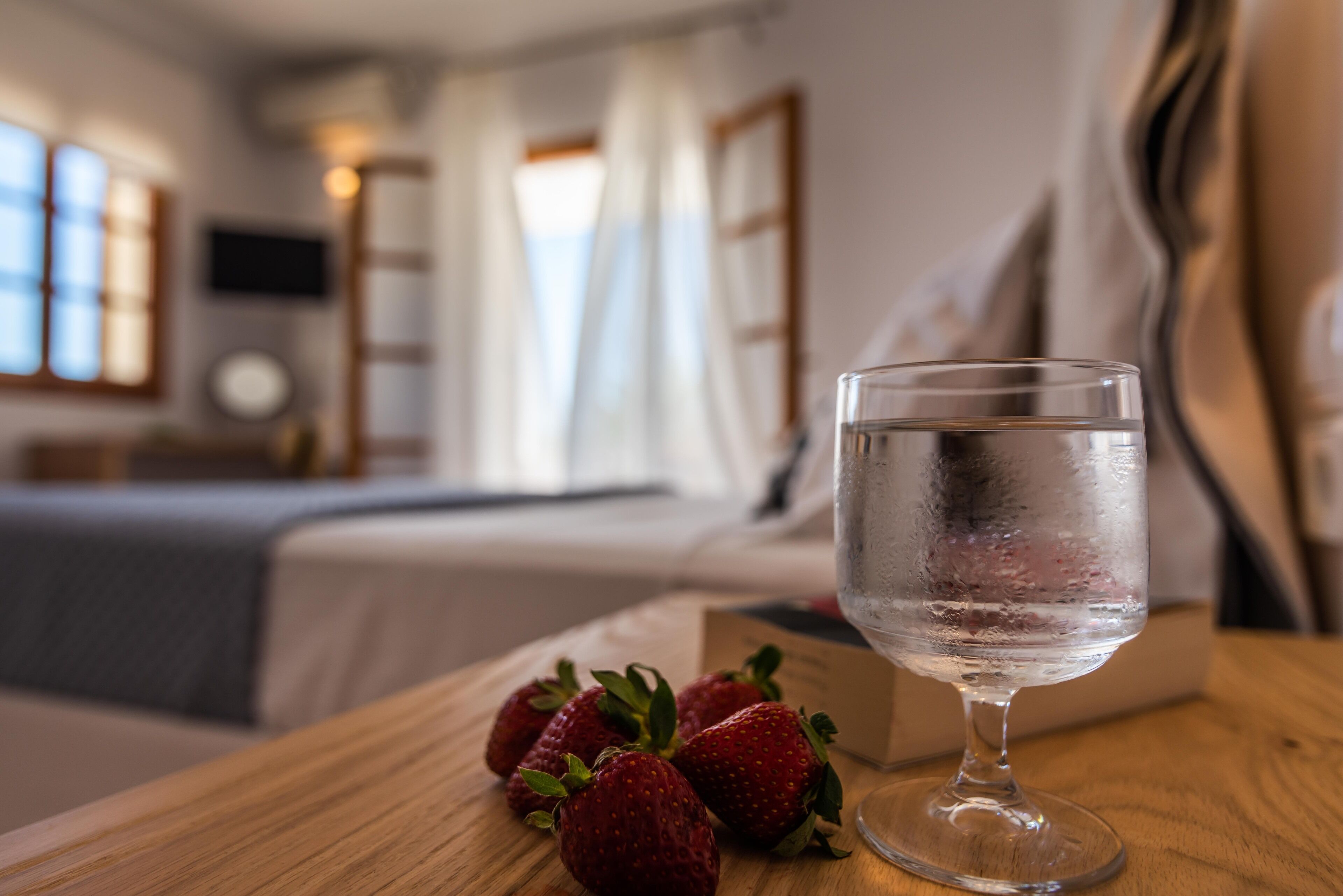 A room with a bed, a bedside table with a glass of water and strawberries, and a TV on the wall.