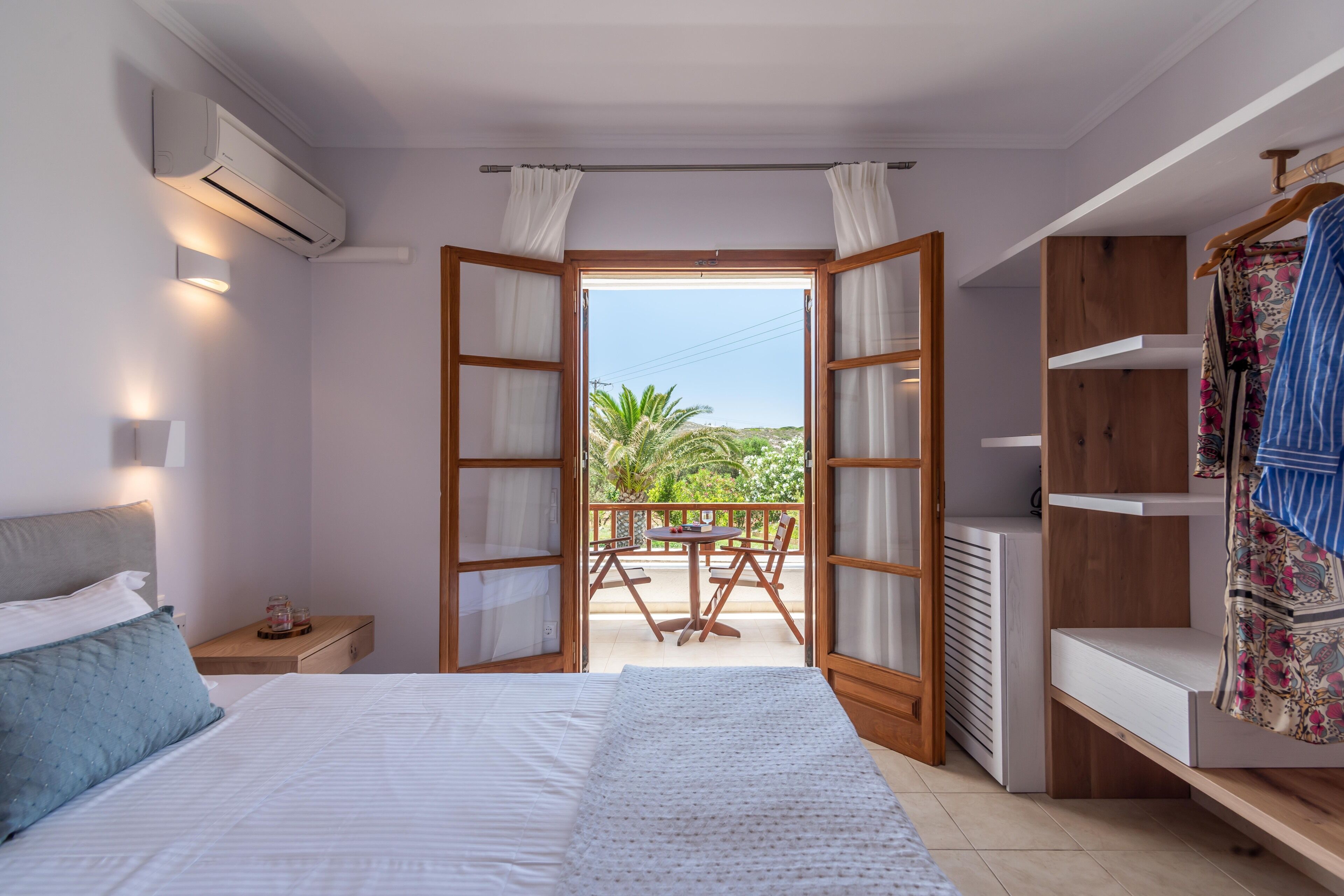 A bedroom with a bed, bedside table, and an open door leading to a balcony with a table and chairs.