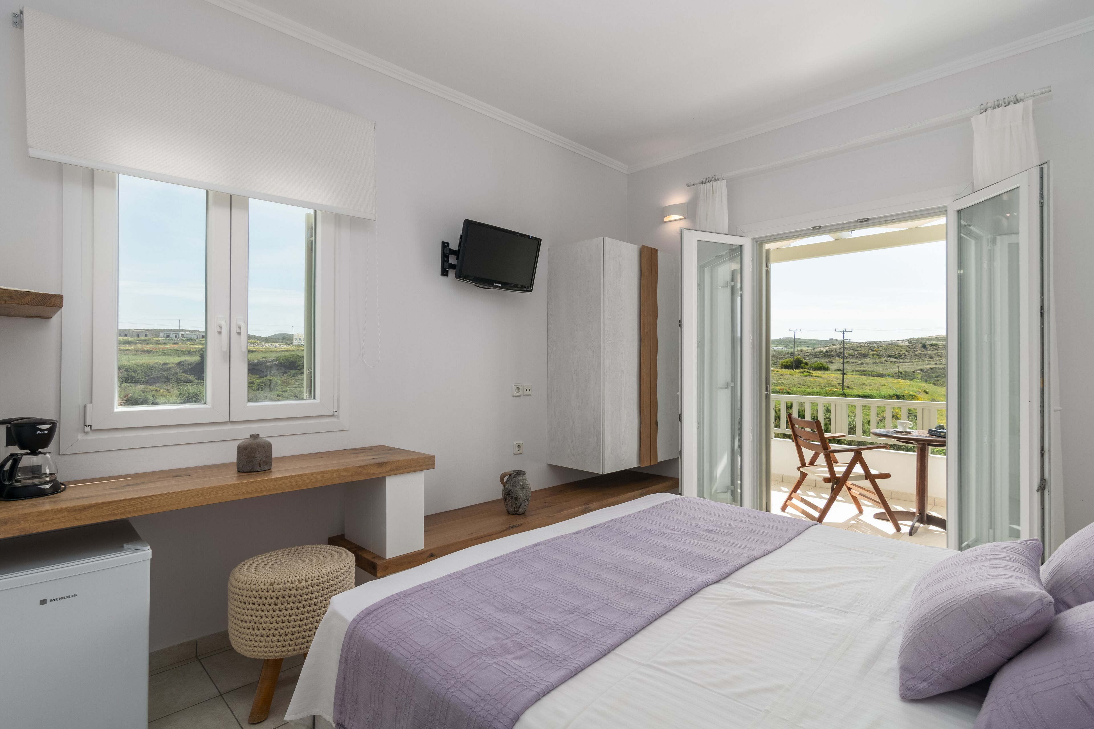 A bedroom with a bed, a desk, a television, and a balcony with a view of the countryside.