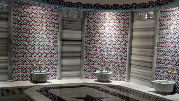 Uma área de spa com uma banheira de hidromassagem central, paredes revestidas com um padrão ondulado e um teto decorativo com motivos florais.