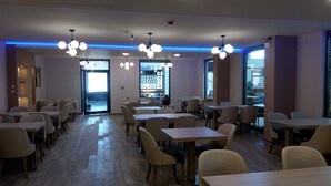 Uma sala de jantar moderna com várias mesas e cadeiras, luminárias no teto e grandes janelas.