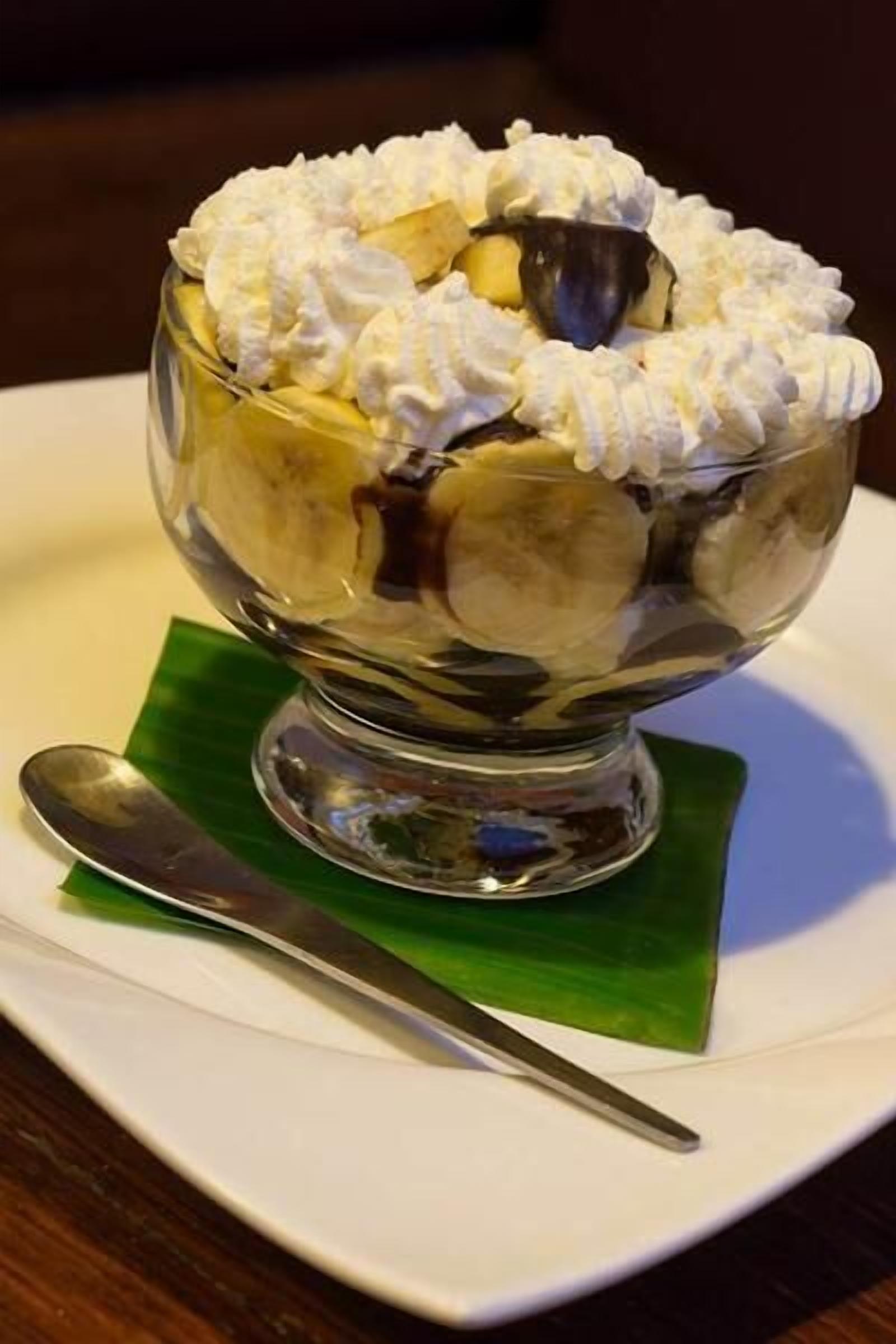 A dessert in a glass bowl with whipped cream on top, placed on a white plate with a green leaf underneath.