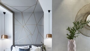 Un dormitori modern amb paret amb dibuixos geomètrics, un llit amb roba de llit blanca i grisosa, una taula auxiliar petita amb un ram i un mirall rodó damunt d'una console.