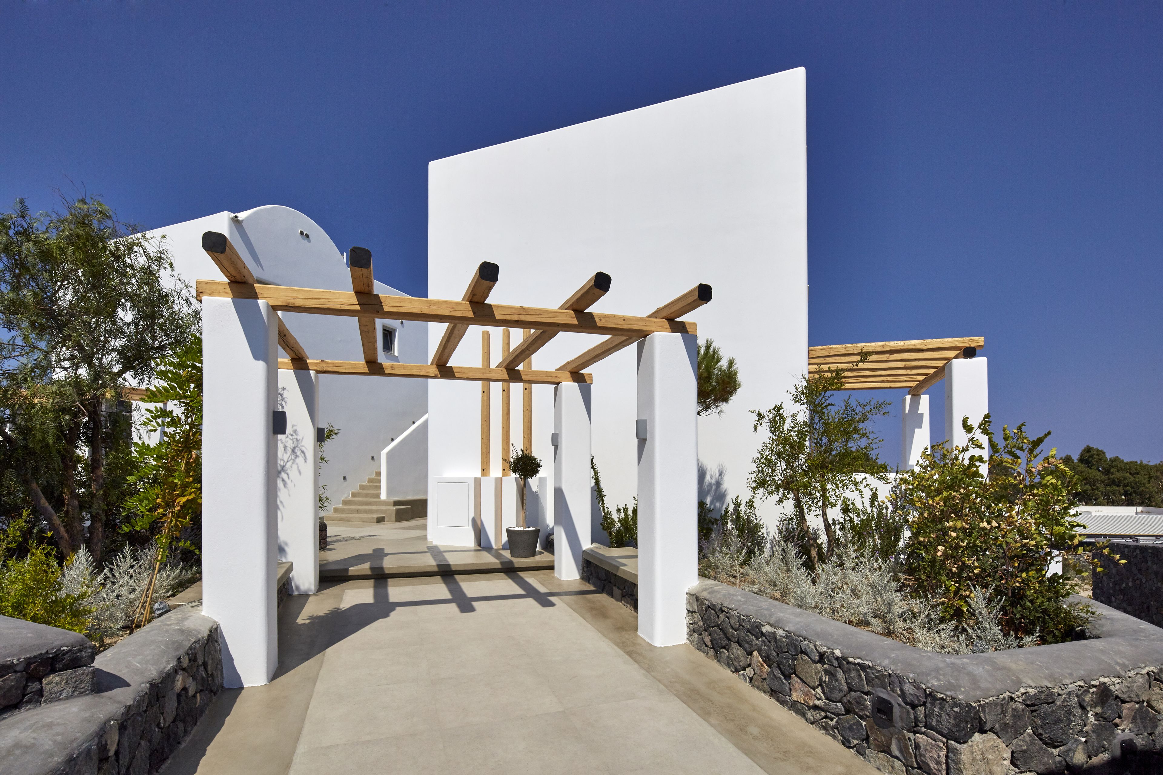 A modern architectural structure with a wooden pergola, stone walls, and a clear sky.