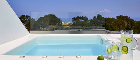 A pool with a clear view of trees and buildings in the background, a glass pitcher with lime slices, and a lime on the poolside.
