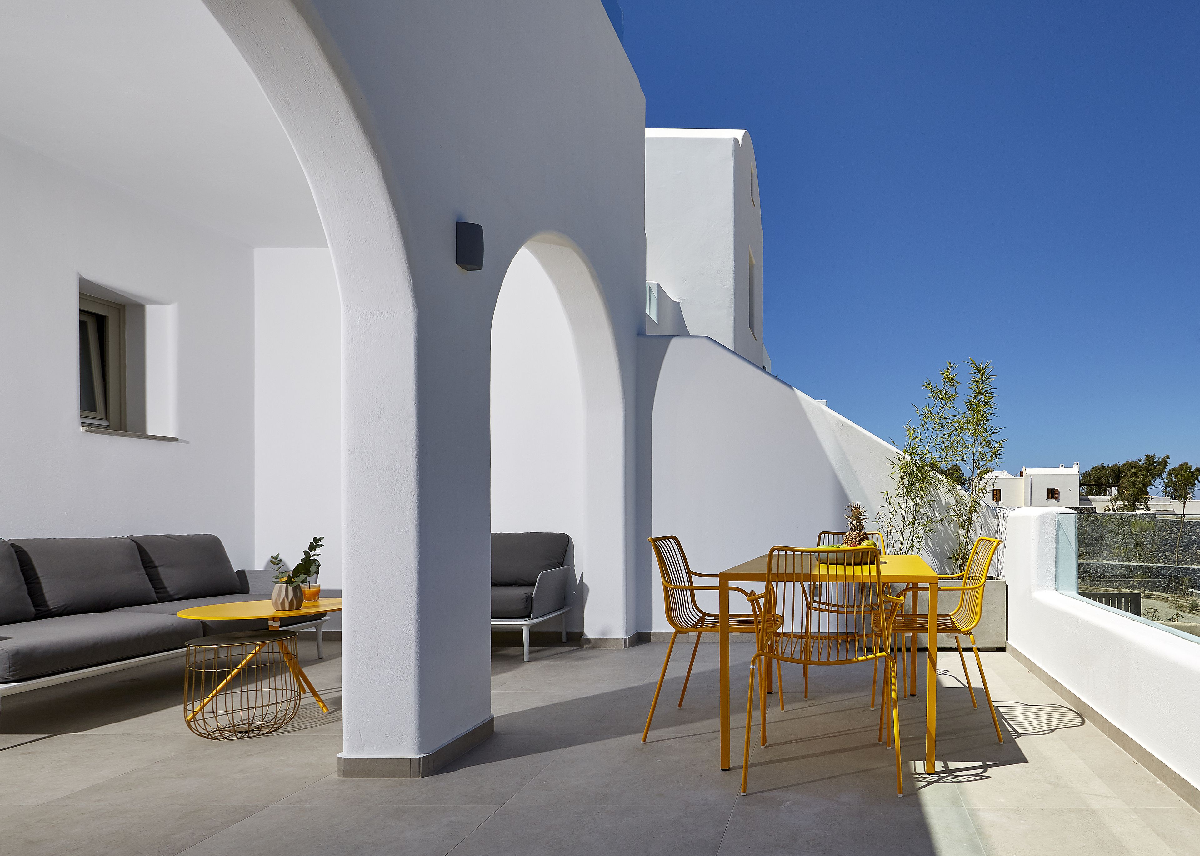 Un espacio moderno al aire libre con sofá, mesa y sillas, rodeado de paredes blancas y arcos, con vistas a un cielo azul despejado.