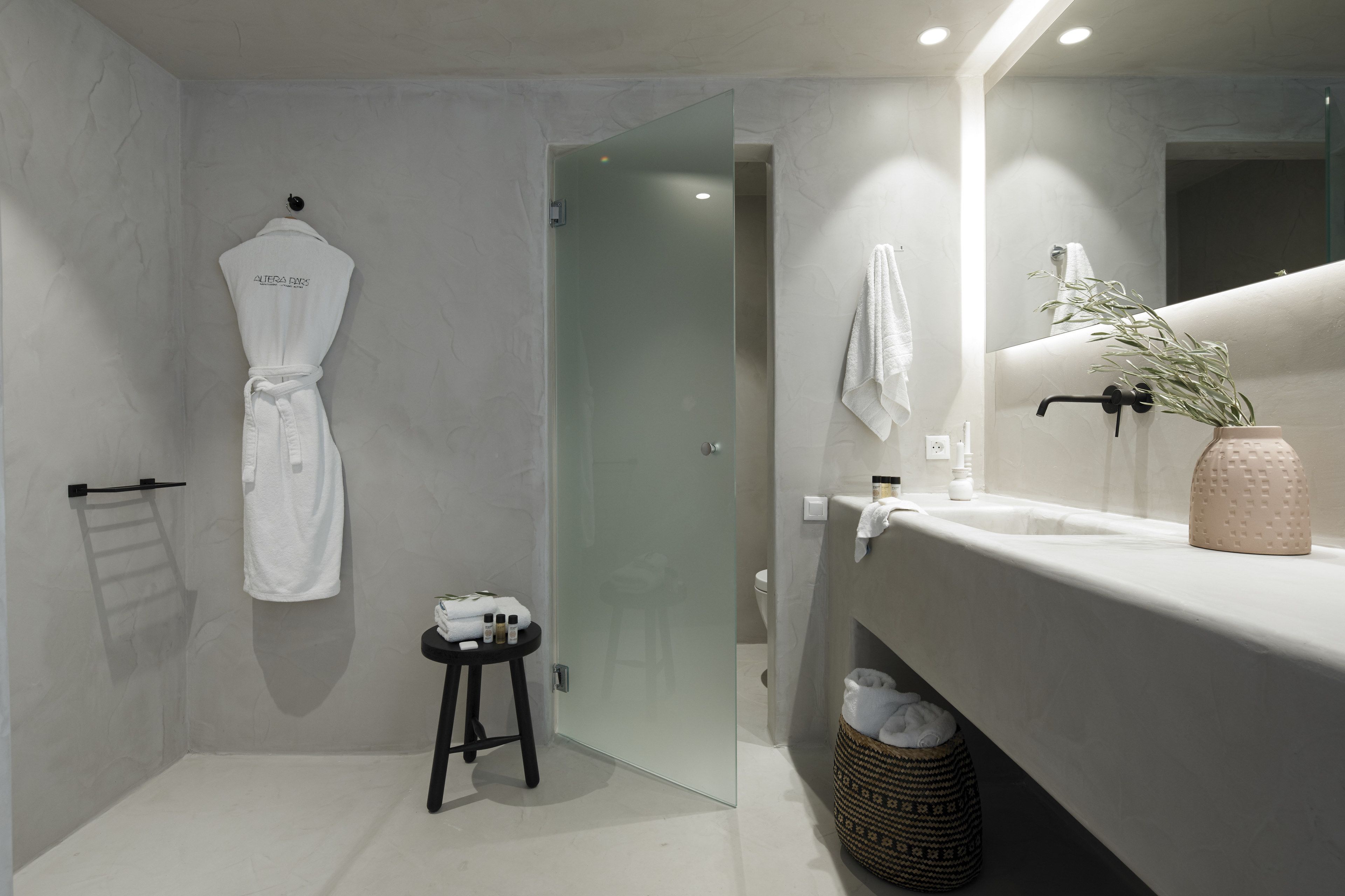 A modern bathroom with a glass shower door, a white robe hanging on a hook, a small stool, and a sink with a vase of greenery.