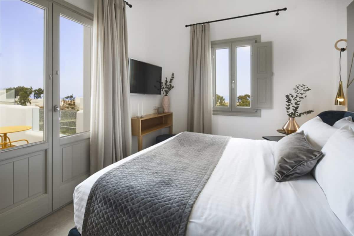 A bedroom with a bed, a TV, a window with curtains, a bedside table, and a view of the outdoors.