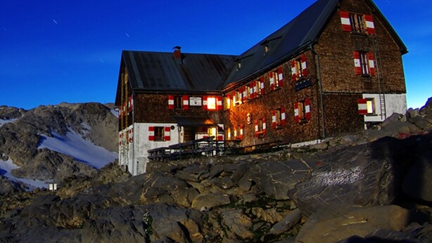 A large, rustic mountain lodge with a steep roof, multiple windows with red shutters, and a stone facade. - Kella-Egg (Brand)