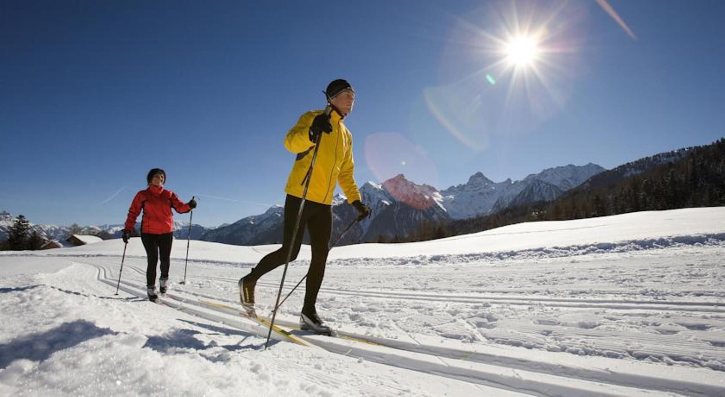 Deux personnes pratiquent le ski de fond sur un sentier enneigé, avec des montagnes en arrière-plan.