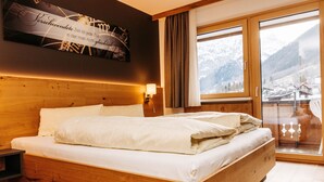 A wooden bed with white bedding in a room with a large window, a wall clock, and a view of a snowy mountain. - Kella-Egg (Brand)