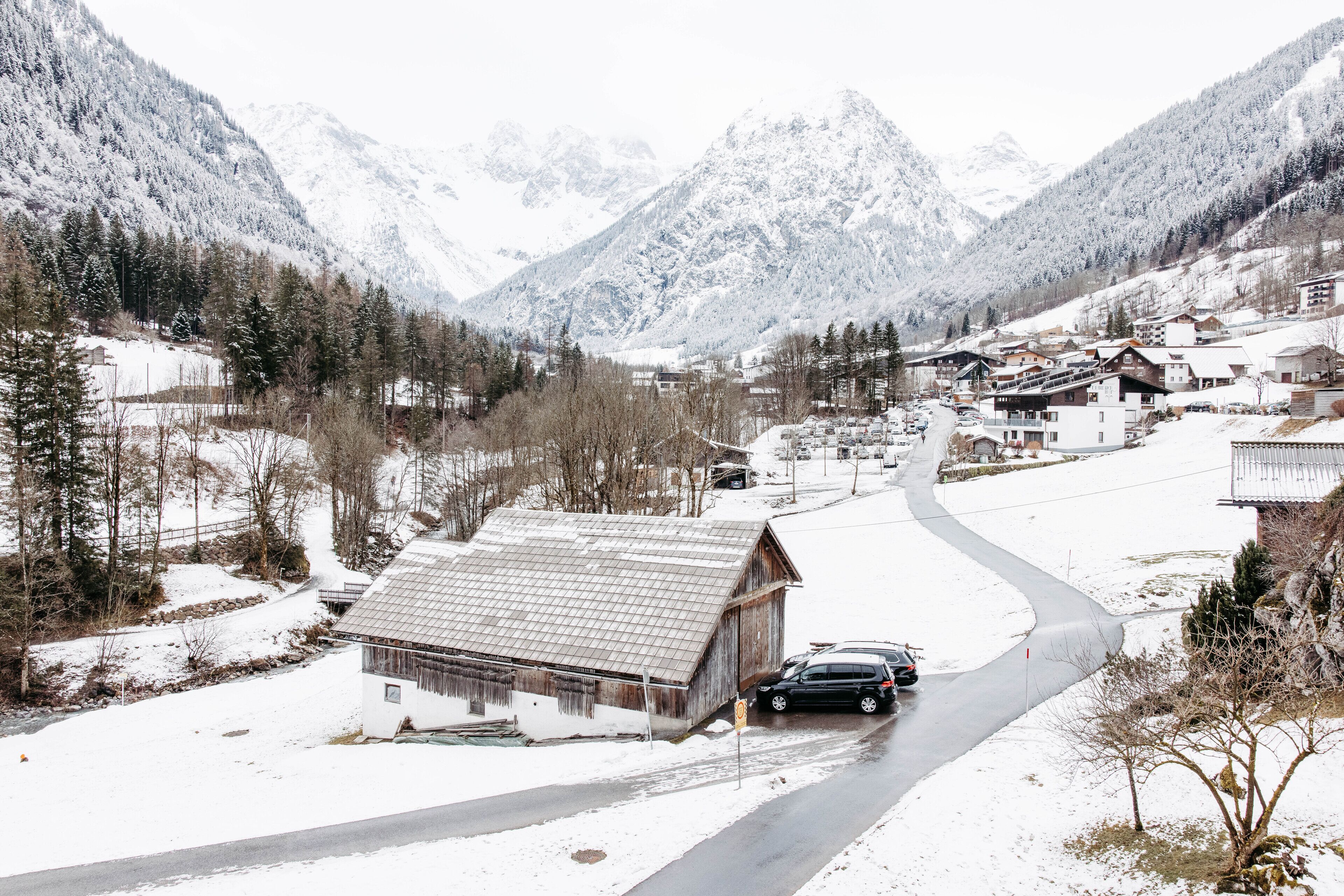 A snow-covered mountain village with a winding road, wooden buildings, and a car parked by the roadside.