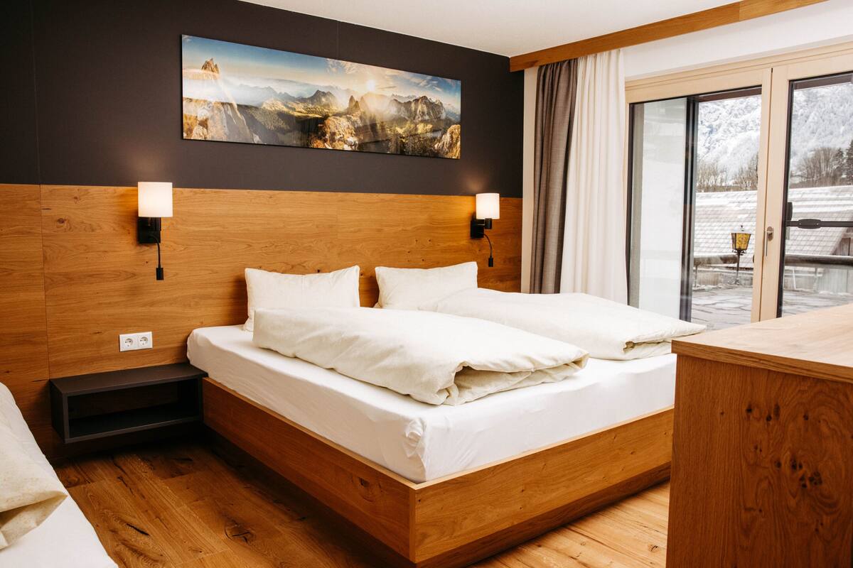 A hotel room with a wooden headboard, two bedside lamps, a large painting of a mountain landscape, and a sliding glass door leading to a balcony.