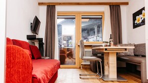 A compact living space with a red sofa, a dining table with chairs, a TV mounted on the wall, and a view of a mountainous landscape through a sliding door. - Kella-Egg (Brand)