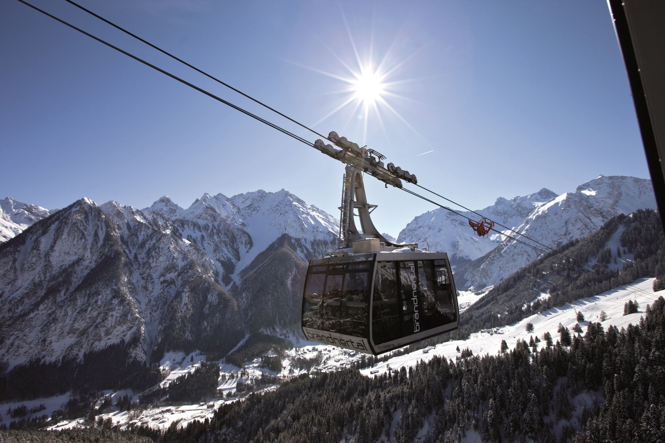 A cable car with the text "Brandneral" in a mountainous landscape with snow-covered peaks and clear skies.