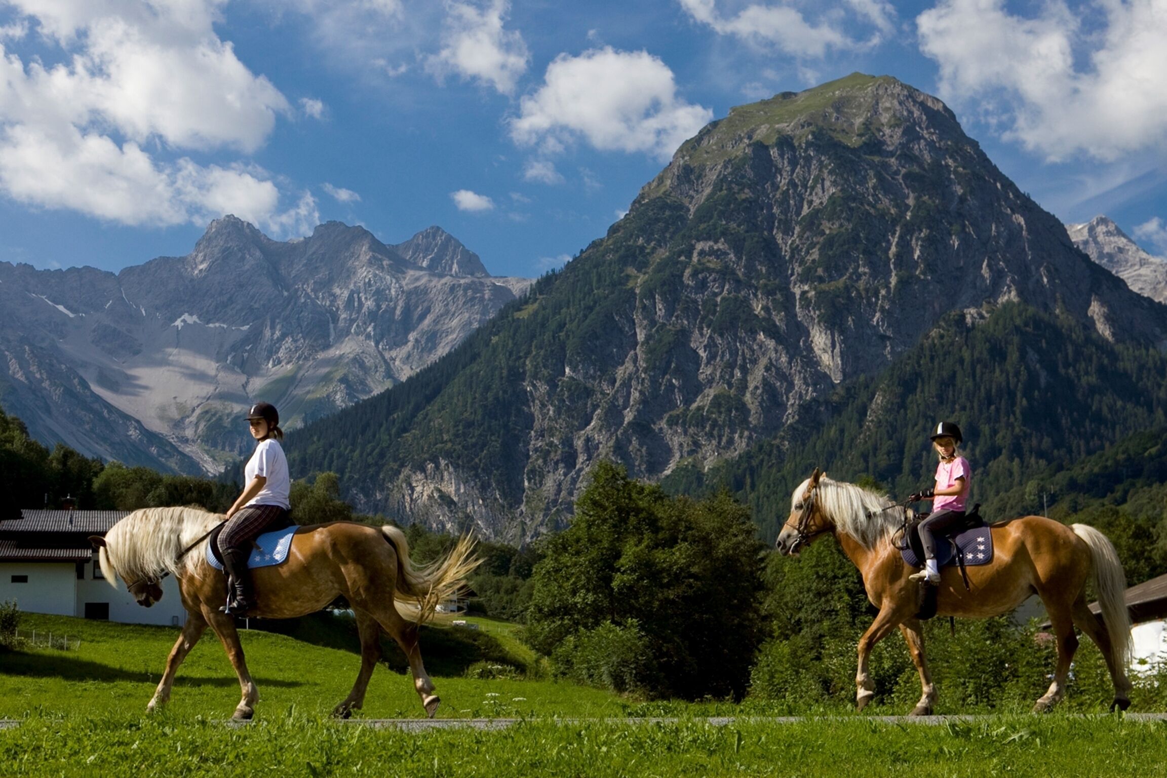 Deux personnes à cheval, évoluant sur un sentier herbeux avec une chaîne de montagnes en arrière-plan.