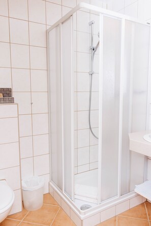 A shower enclosure with a glass door, a white toilet, and a trash bin in a bathroom with white tiled walls and floor. - Kella-Egg (Brand)