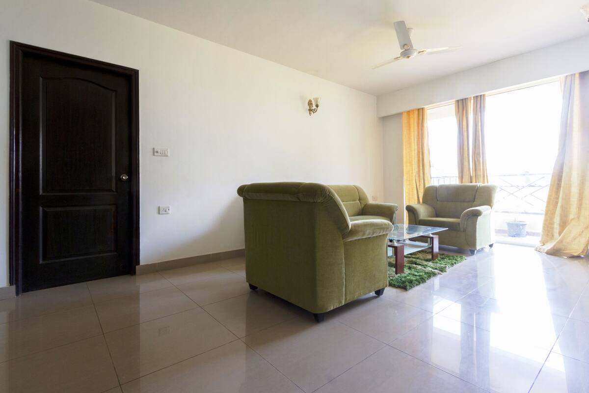Un salón espacioso con una puerta de madera oscura, un sillón verde, un sillón beige y una mesita.