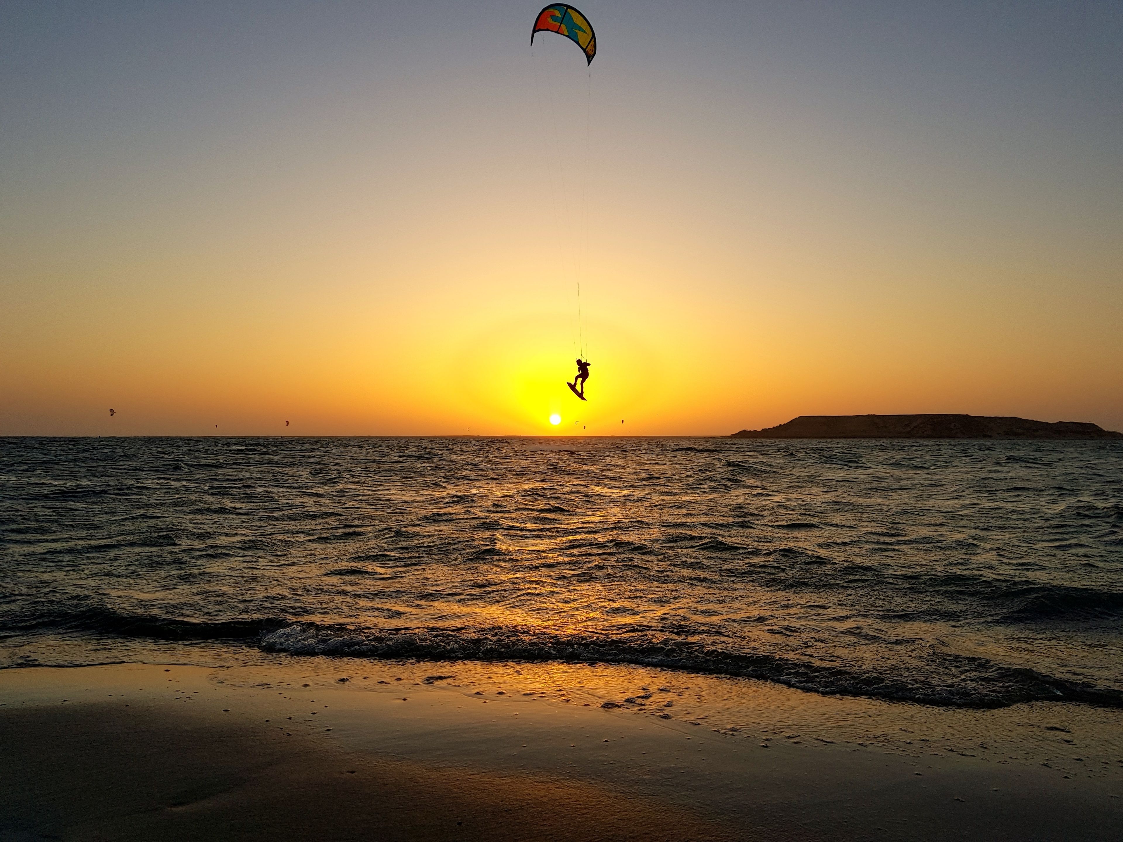 Une plage au coucher du soleil, avec des vagues et un pratiquant de kitesurf.