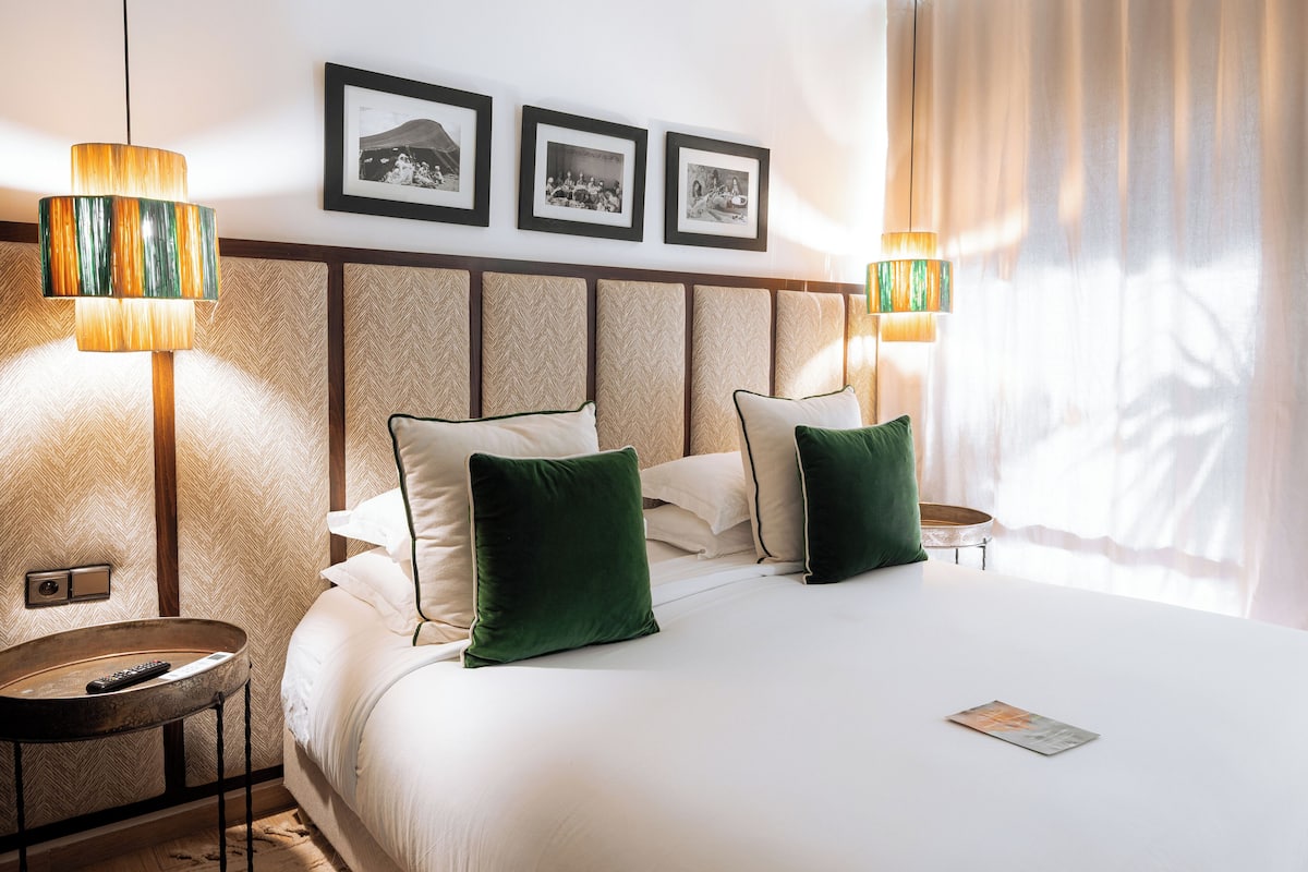 Habitación de hotel con una cama grande, dos lámparas de noche, una mesita pequeña y tres cuadros en la pared.