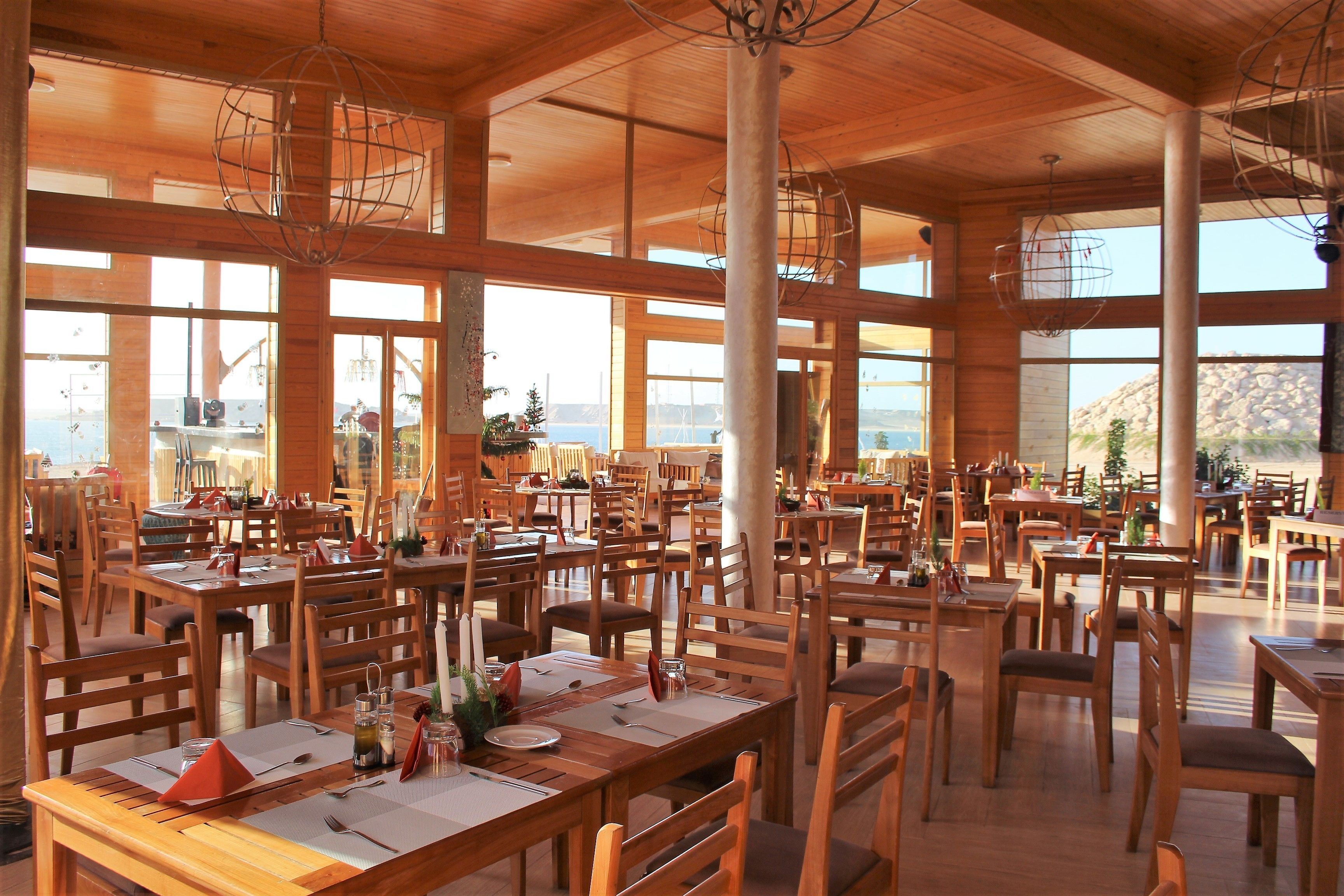 A spacious restaurant with wooden tables and chairs, large windows, and a view of the sea.