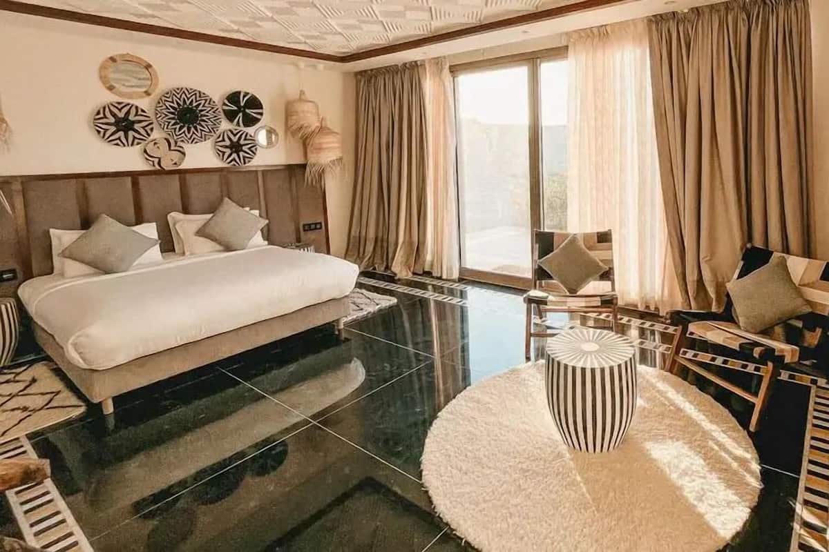 Una habitación de hotel moderna con una cama grande, una mesa de centro redonda y vistas al exterior a través de amplios ventanales.