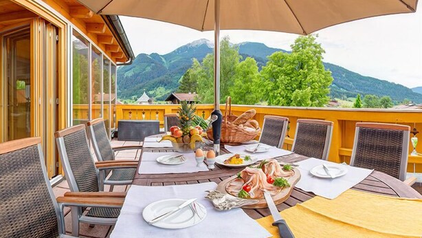 An outdoor dining area with a table set for two, surrounded by wicker chairs, a large umbrella, and a scenic mountain view. - Landvilla Romantika (Lermoos)
