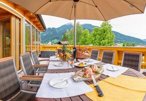Un espacio de comedor al aire libre con una mesa preparada para dos personas, rodeado de sillas de mimbre, un paraguas grande y una vista panorámica a las montañas.