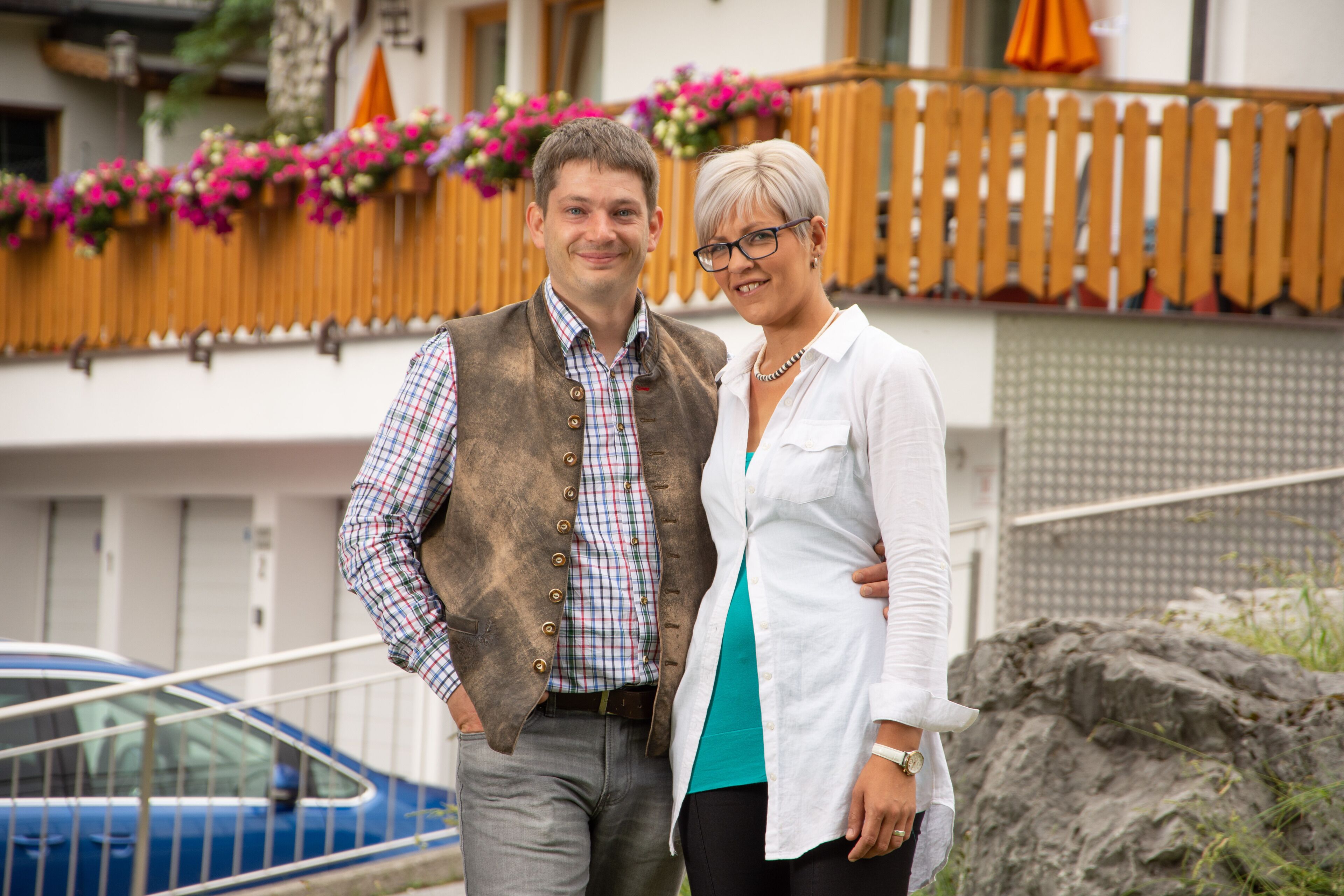 A man and a woman standing in front of a building with a wooden fence and flowers.