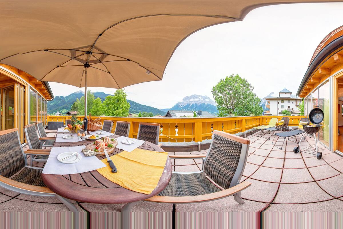 A dining area with a table set for two, surrounded by chairs, and a large umbrella providing shade.