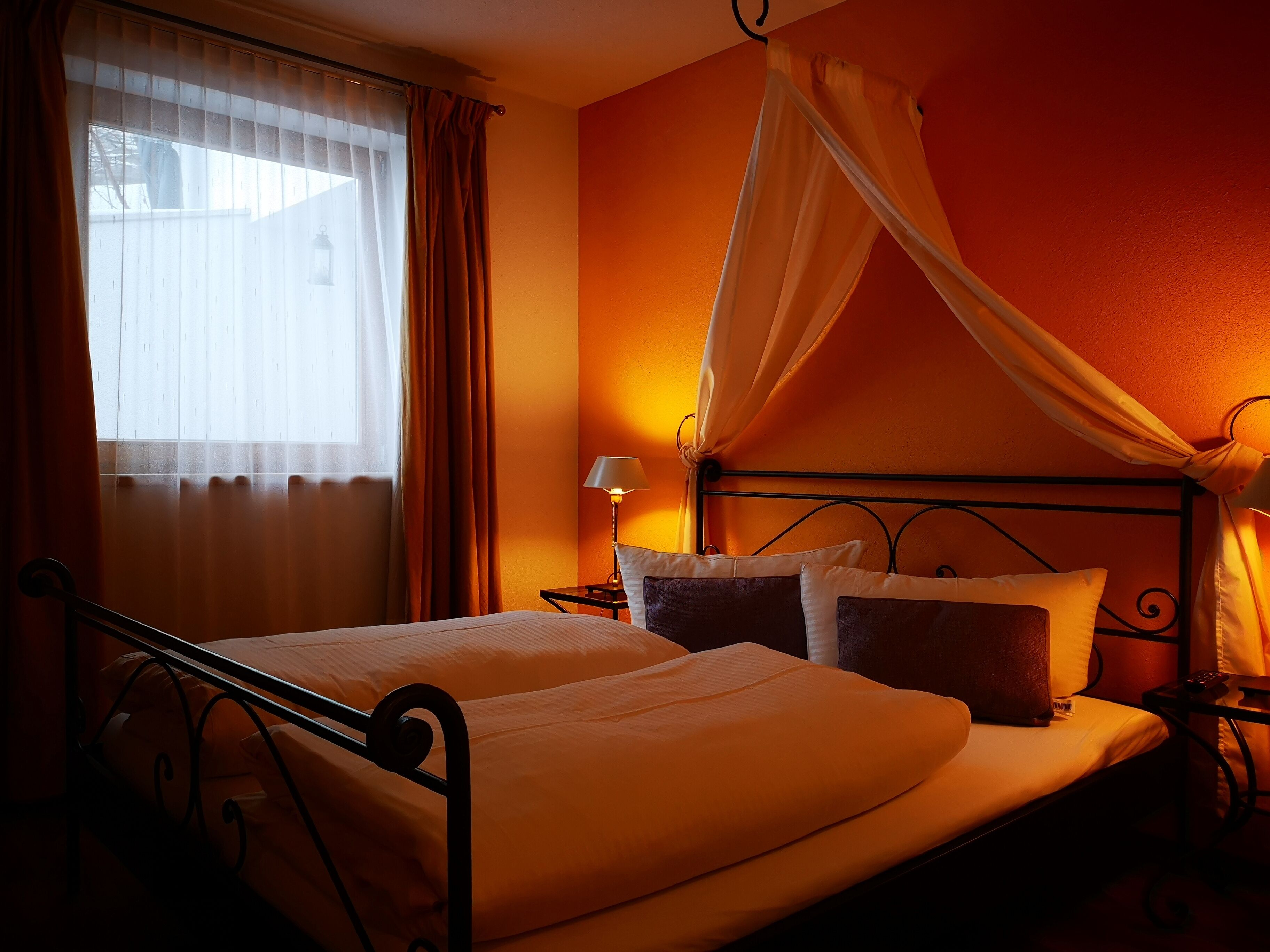 Ein Doppelbett mit Baldachin, ein Nachttisch mit Lampe und ein Fenster mit Vorhängen.