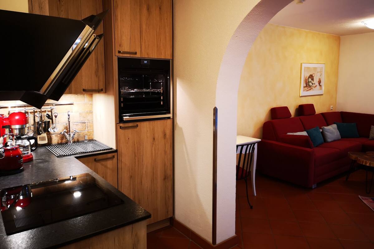A kitchen with wooden cabinets, a built-in oven, and a red mixer. There is a red couch in a living area with a painting on the wall.