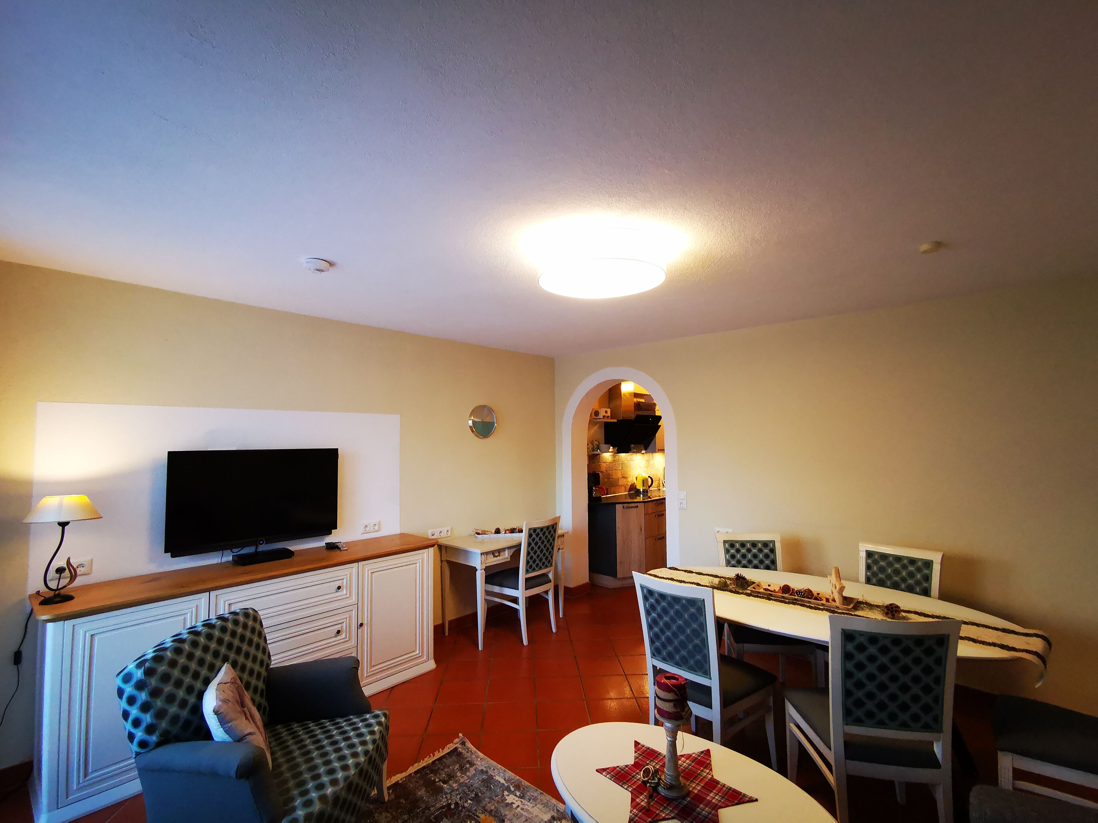 A living room with a TV, dining table, chairs, and a kitchen visible through an archway.