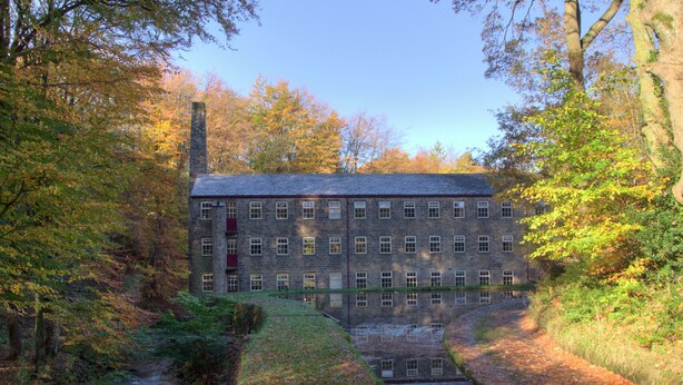 A stone building with multiple windows, surrounded by trees and a small water channel. - Hewenden Mill Holiday Homes (Bradford)