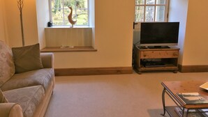 A living room with a sofa, a lamp, a television on a wooden stand, and a window with a decorative bird. - Hewenden Mill Holiday Homes (Bradford)