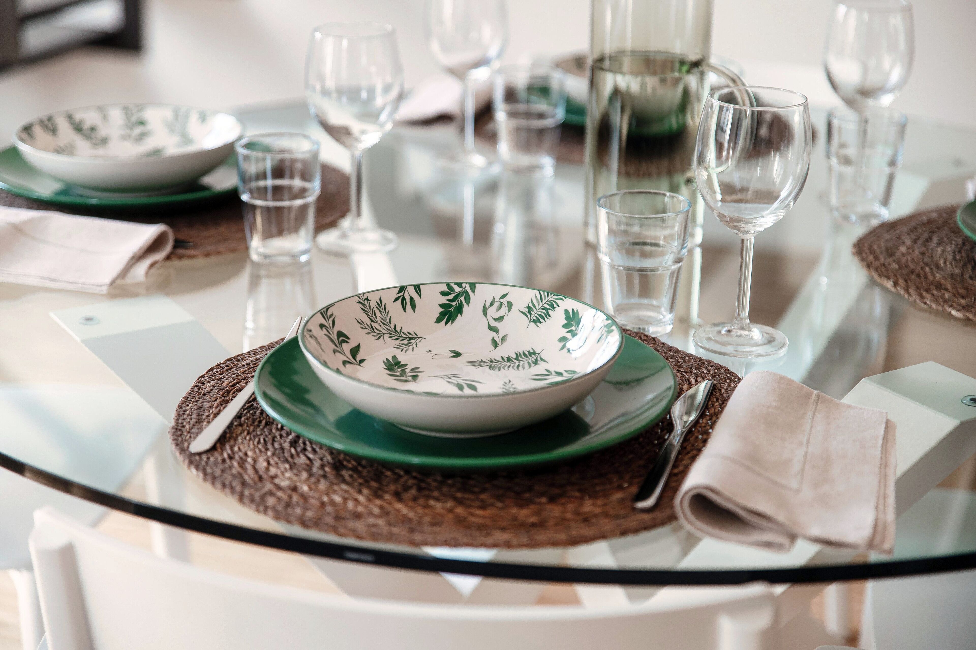 Una mesa redonda de cristal con un posavasos verde, un cuenco con estampado de hojas y una servilleta doblada.