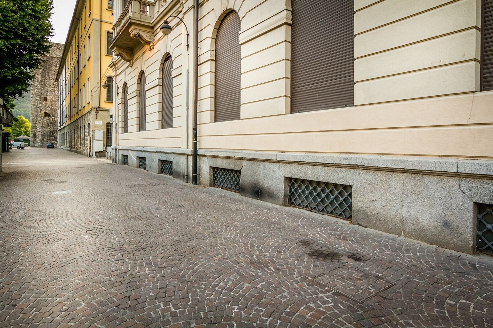 Une rue pavée bordée de bâtiments historiques, dont un immeuble jaune doté de fenêtres en arc de cercle et un mur de pierre surmonté d’une grille métallique.