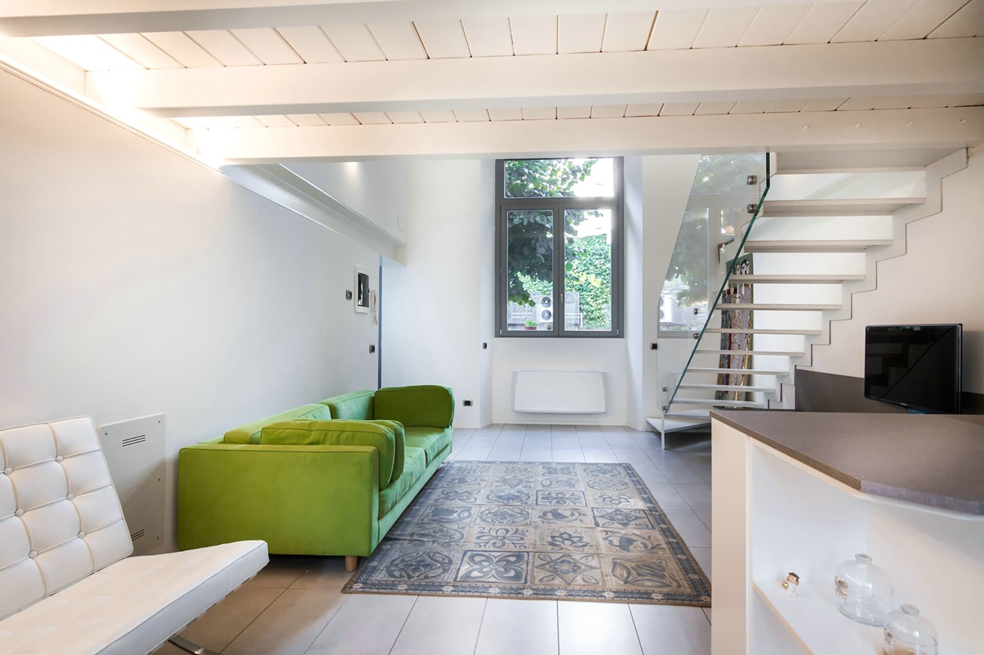 Un salon moderne avec un canapé vert, un divan blanc, un tapis à motifs et un escalier menant à un niveau supérieur.