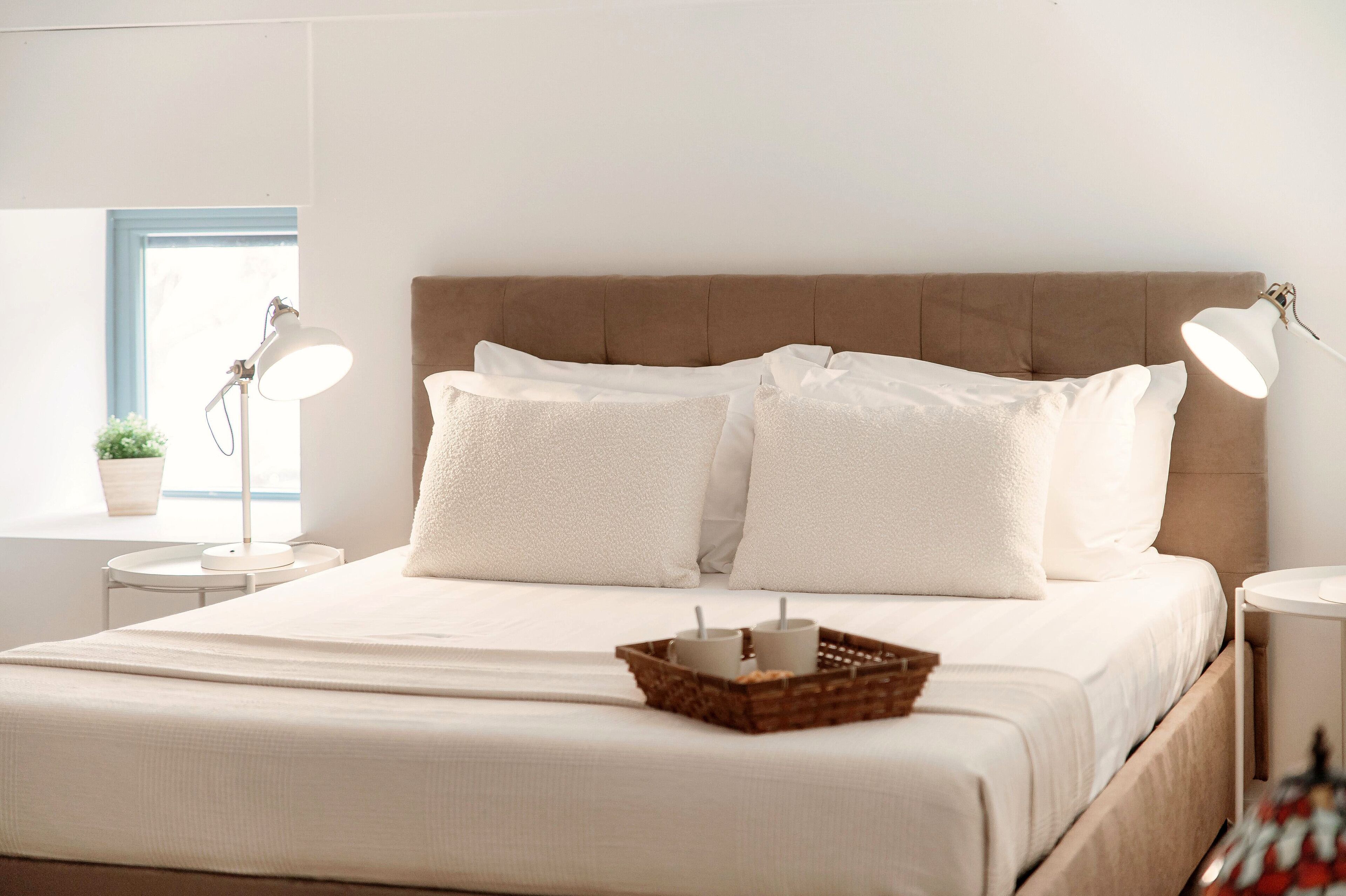 A neatly made bed with white linens, two bedside lamps, and a small bedside table with a plant and a tray of candles.