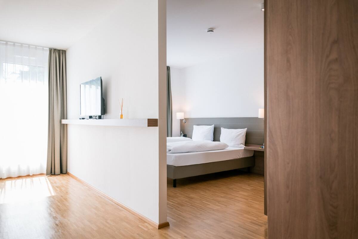 A modern hotel room with a wooden floor, a bed with white linens, a nightstand, a TV on a shelf, and a window with curtains.
